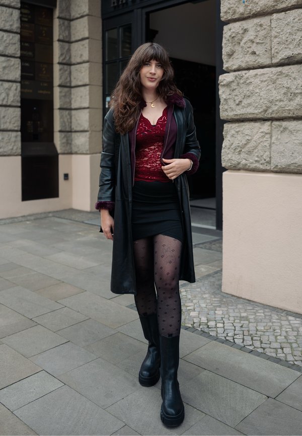 Une femme se tient devant un bâtiment en pierre. Elle porte un long manteau noir, un haut rouge en dentelle, une jupe noire, des collants à motifs et des bottes.