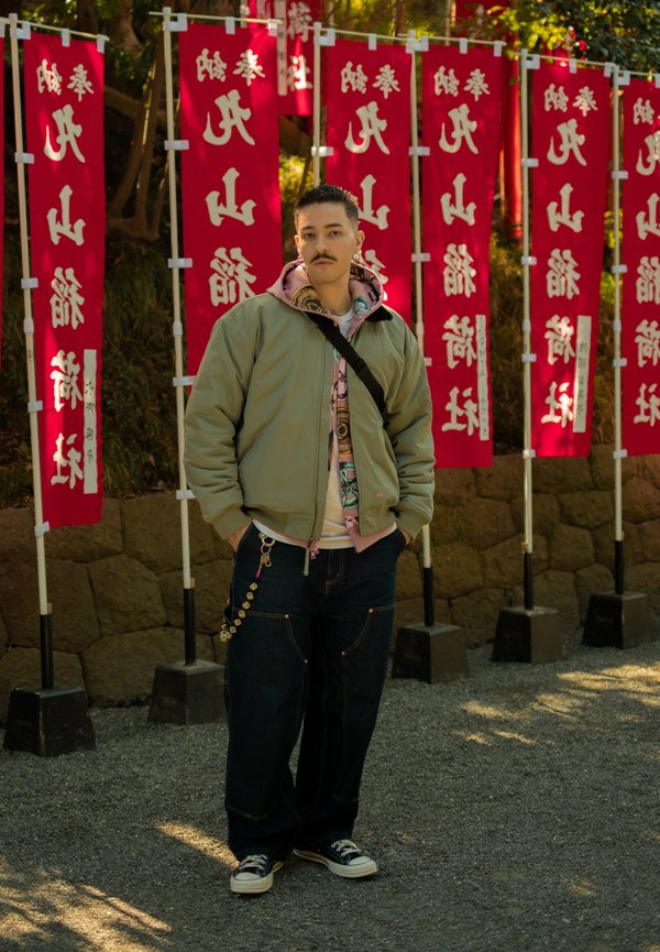 Mand med moustache iført grøn jakke og jeans, står foran røde bannere med hvide japanske tegn udendørs.