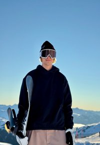 Snowboarder che indossa un felpa con cappuccio nera, pantaloni chiari e guanti bianchi, mentre tiene una tavola da snowboard con attacchi neri e rossi.