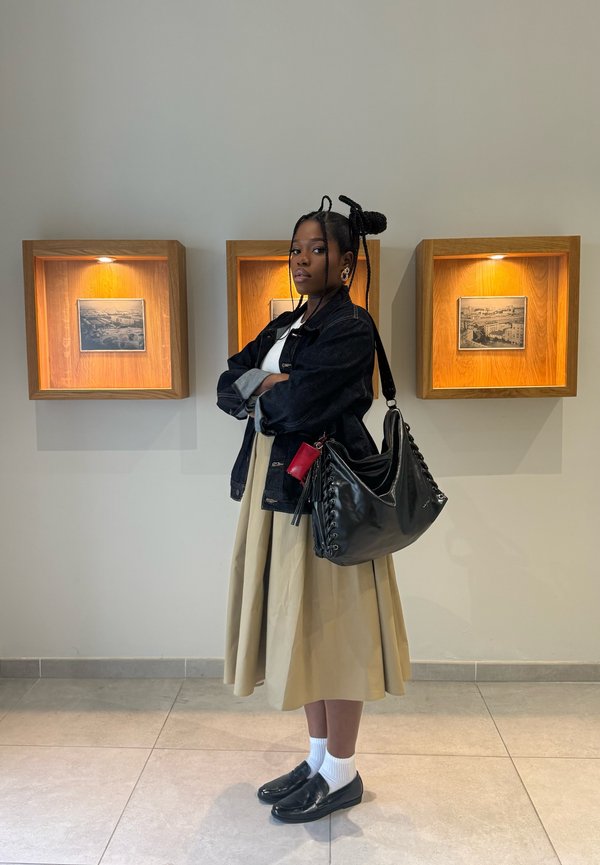 Młoda kobieta z warkoczami stoi ze skrzyżowanymi ramionami przed trzema drewnianymi skrzyniami wystawowymi z podświetlonymi czarno-białymi fotografiami.