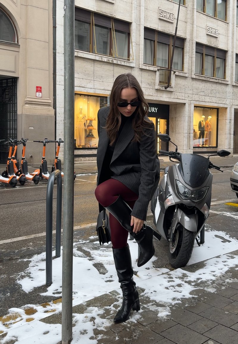 Donna con occhiali da sole che sistema stivali neri alti fino al ginocchio su un marciapiede innevato accanto a uno scooter parcheggiato e una fila di monopattini elettrici arancioni.