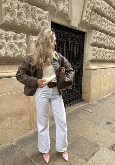 Chaqueta de cuero marrón, suéter crema, pantalones anchos blancos, zapatos de punta rosa, sosteniendo un bolso marrón con estampado; fondo de pared de piedra.