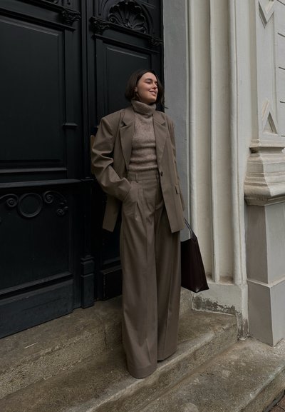 Blazer beige de corte oversized y pantalones anchos a juego sobre un ligero cuello de tortuga. Los accesorios incluyen un bolso marrón oscuro. De pie junto a una puerta.