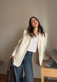 Hvit oversized blazer med slagkrage, brukt over en hvit topp og lyseblå jeans. Brun håndveske henger fra den ene armen; moderne laptop på bordet.