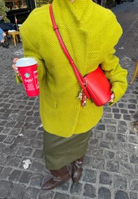 Pull vert surdimensionné en tricot avec un col montant, associé à une jupe en cuir marron et des bottes marron montantes jusqu'aux genoux. Tenant une tasse Starbucks rouge et un sac bandoulière rouge.