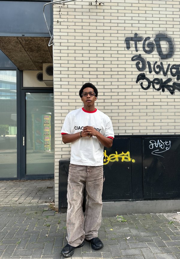 Hvit t-skjorte med røde detaljer og tekst, lyse beige bukser, og svarte sko. Står mot en murvegg med graffiti.