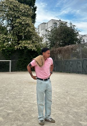 Camisa polo rosa claro, jeans azul claro, suéter beige caído sobre el hombro, zapatillas verdes con rayas blancas y un reloj en la muñeca.