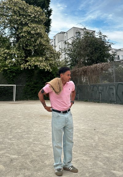 Camisa polo rosa claro, jeans azul claro, suéter beige caído sobre el hombro, zapatillas verdes con rayas blancas y un reloj en la muñeca.
