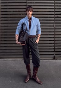 Cardigan bleu clair à fermeture éclair, chemise marron à pois, pantalon gris fuselé, bottes en cuir marron et sac cabas noir, sur fond à lattes.