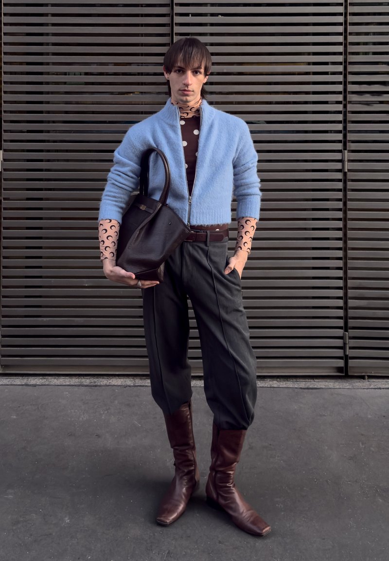 Cardigan bleu clair à fermeture éclair, chemise marron à pois, pantalon gris fuselé, bottes en cuir marron et sac cabas noir, sur fond à lattes.