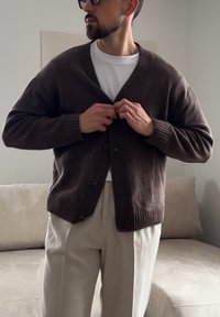 Homme avec barbe et lunettes de soleil boutonnant un cardigan brun foncé sur une chemise blanche, portant un pantalon beige clair, debout près d'un canapé beige.