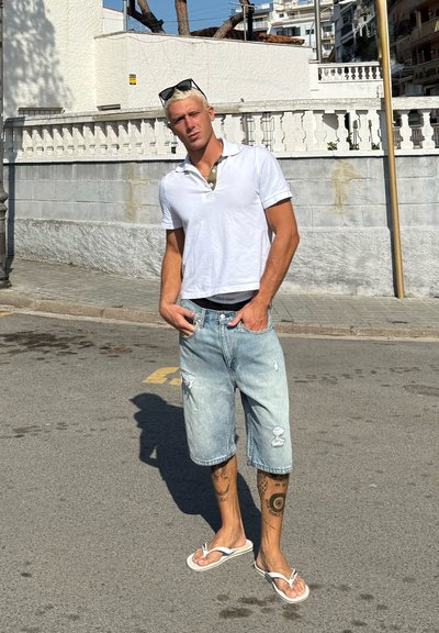 Camisa polo blanca sobre un cuello verde, pantalones cortos de mezclilla desgastados de color azul claro y chanclas blancas. Tatuajes visibles en brazos y piernas.