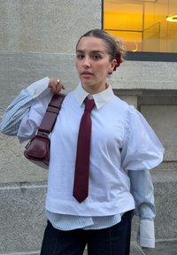 Camicia bianca oversize con maniche a sbuffo, cravatta bordeaux e camicia a righe blu sotto. Pantaloni in denim scuro e una piccola borsa bordeaux.