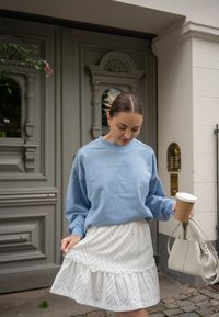 Pullover in hellblau mit besticktem Text; weißer Rock mit Rüschen aus Lochspitze; hält eine Kaffeetasse; beiger Handtasche. Der Hintergrund zeigt verzierte Türen.