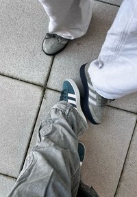 Two pairs of shoes on gray pavement: one green with white stripes, another gray with dark soles. Both have a casual design and textured surfaces.