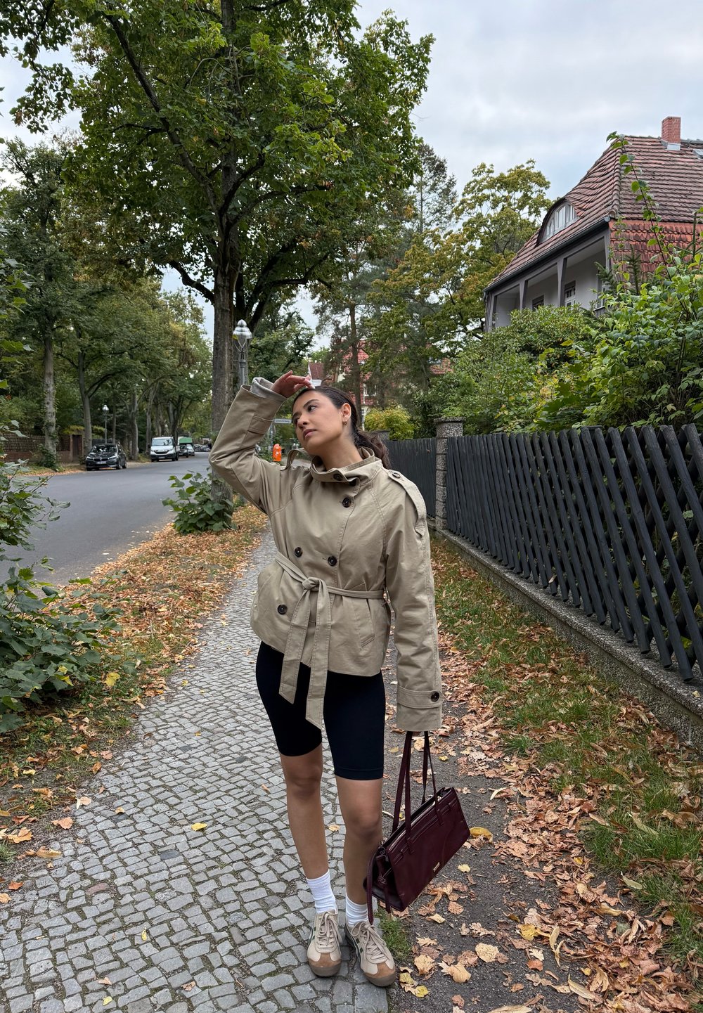 Giacca beige con cintura e bottoni doppi, abbinata a pantaloni ciclisti neri. Scarpe marroni e una borsa bordeaux completano il look.