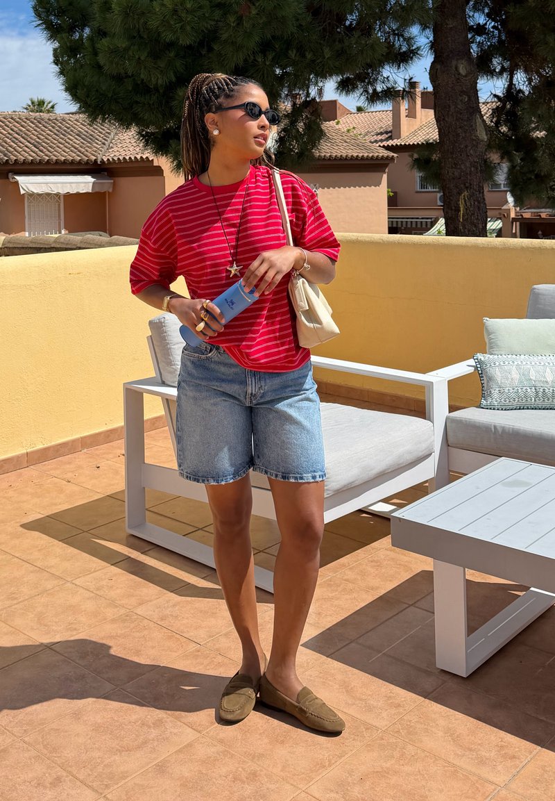 Femme portant des lunettes de soleil, chemise rouge à rayures, short en jean, tenant une bouteille bleue, debout sur une terrasse ensoleillée avec mobilier extérieur.