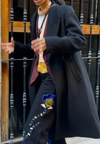 Zwarte wollen jas met gestructureerde revers, over een bordeaux rode vest en witte shirt. Zwarte broek met kleurrijke bloemembroidery. Beige gestreepte sjaal.