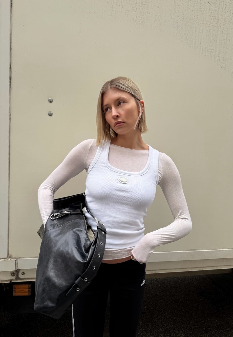 Junge Frau mit kurzen blonden Haaren, die geschichtete weiße Oberteile und schwarze Hosen trägt und eine große schwarze Ledertasche über der Schulter hat.