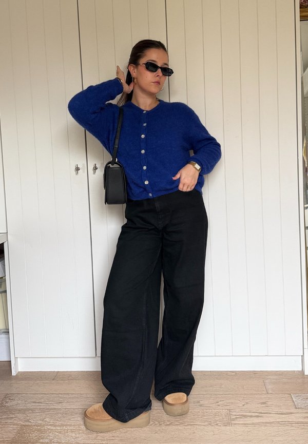 Modrý vlněný kardigan s zapínáním na knoflíky, kombinovaný s širokými černými kalhotami a béžovými platformovými botami. Černé obdélníkové sluneční brýle a malá taška.