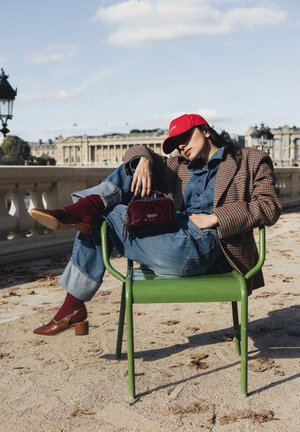 Mujer con blazer a cuadros, mono vaquero, gorra roja y zapatos marrones recostada en una silla de metal verde al aire libre con un edificio histórico de fondo.
