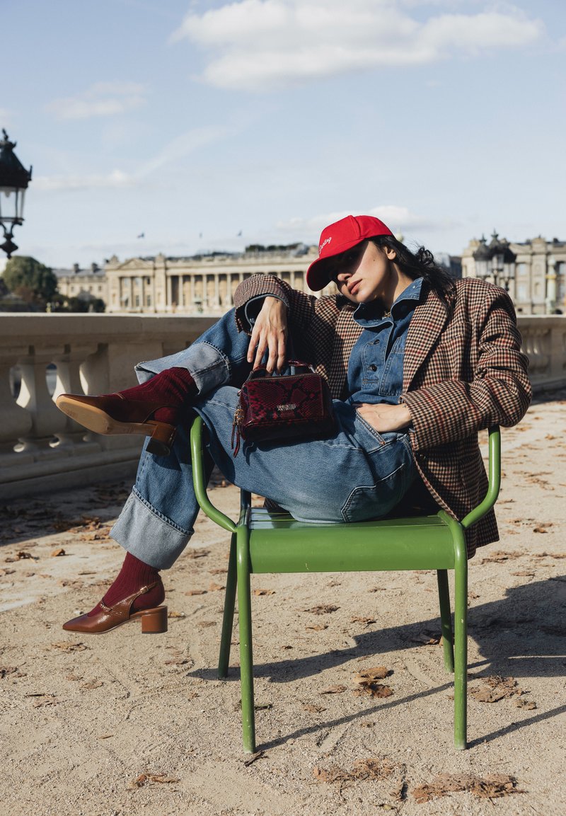 Femme en blazer à carreaux, combinaison en jean, casquette rouge et chaussures brunes, allongée sur une chaise en métal verte en plein air avec un bâtiment historique en arrière-plan.