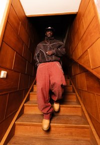 Άντρας που φορά καφέ μπουφάν, καπέλο, κόκκινο καρό παντελόνι και ταμπά μπότες, κατεβαίνει ξύλινη σκάλα με επενδεδυμένους τοίχους και κουπαστές.