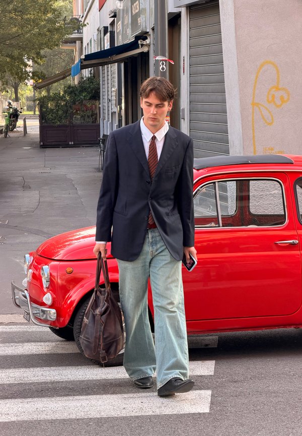 Un joven con traje oscuro y vaqueros cruza la calle delante de un coche clásico rojo, llevando un bolso de cuero y un teléfono.