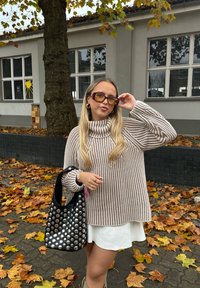 Gestreepte beige coltrui, witte rok, zwarte tas met zilveren studs; herfstbladeren en een boom op de achtergrond.