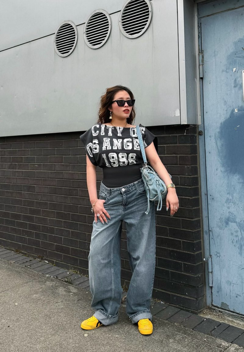 Femme portant un haut noir "City of Los Angeles 1998", un jean bleu ample, des chaussures jaunes, des lunettes de soleil et un sac à bandoulière bleu, debout à l'extérieur.
