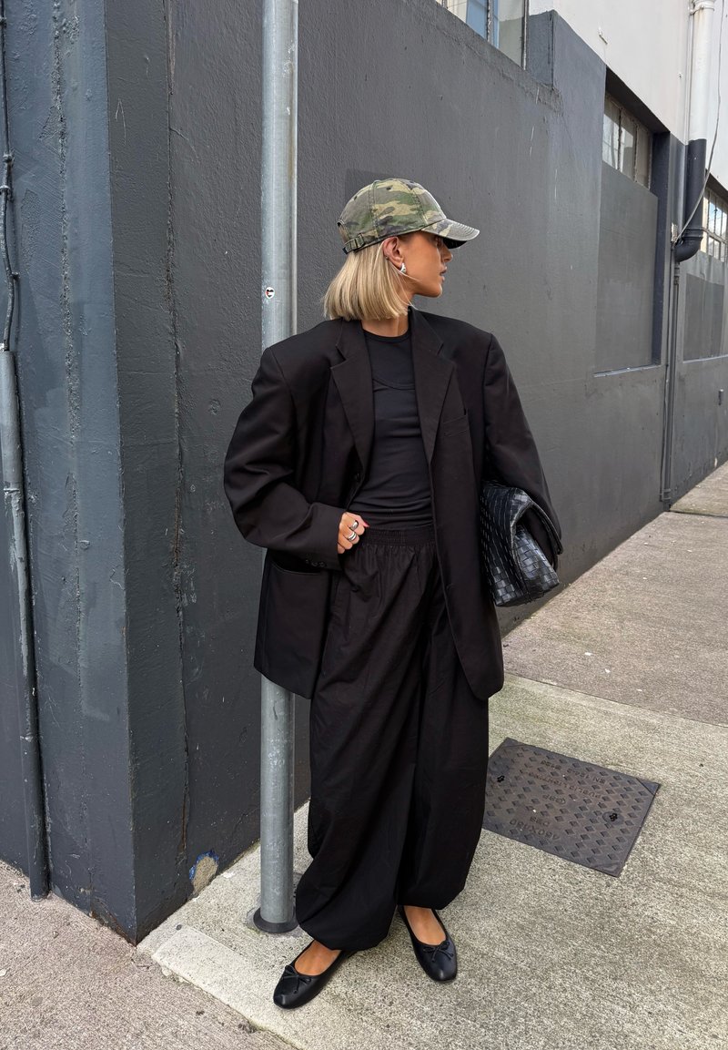 Blazer oversized preto sobre uma blusa preta e calças largas. Os acessórios incluem um boné camuflado e uma carteira preta texturizada. Sapatos pretos planos.