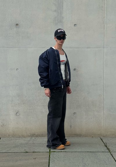 Chaqueta de piloto navy, camiseta blanca sin mangas con logo rojo, jeans negros de pierna ancha, zapatos color beige, gorra negra y gafas de sol, frente a un fondo de hormigón.