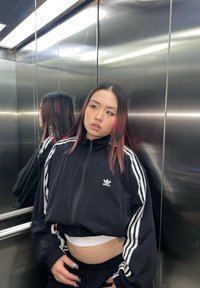 Casaco preto cropped com riscas brancas, usado sobre uma blusa branca; refletido num elevador metálico. Calças pretas e cabelo comprido com mechas rosas.