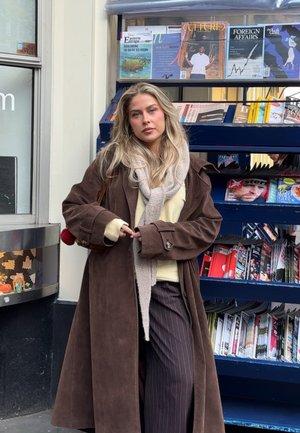Mujer rubia con abrigo largo marrón, bufanda beige y pantalones a rayas, de pie frente a un quiosco lleno de revistas y periódicos.