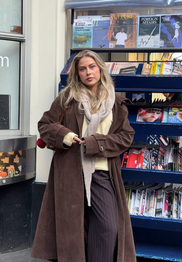 Blondynka w brązowym długim płaszczu, beżowym szalu i pasiastych spodniach stoi przed kioskiem z magazynami i gazetami.