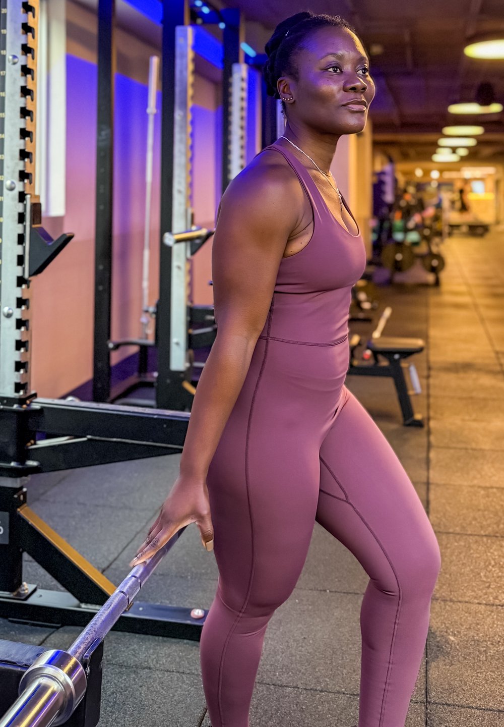 Femme en tenue de sport violette tenant une barre d'haltérophilie derrière son dos dans une salle de sport avec du matériel de musculation et un éclairage chaud.