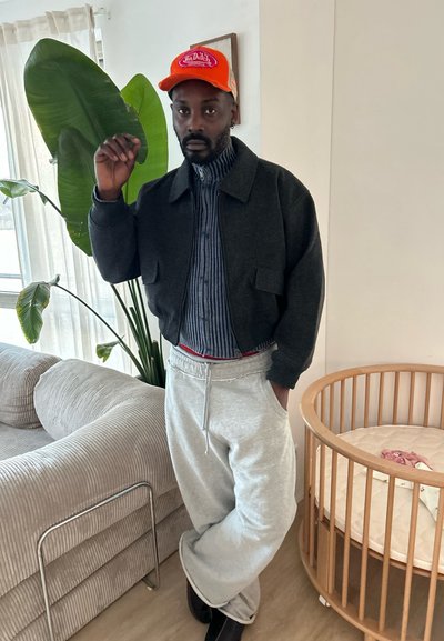 Chaqueta de lana negra sobre una camisa de rayas azules, pantalones de chándal grises y una gorra naranja brillante. De pie al lado de una planta en maceta y una cuna circular.