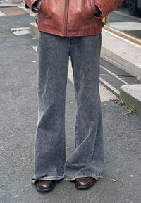 Personne portant un jean gris délavé à jambes larges couvrant des chaussures marron foncé, les mains dans les poches d'une veste en cuir marron, debout sur le trottoir.