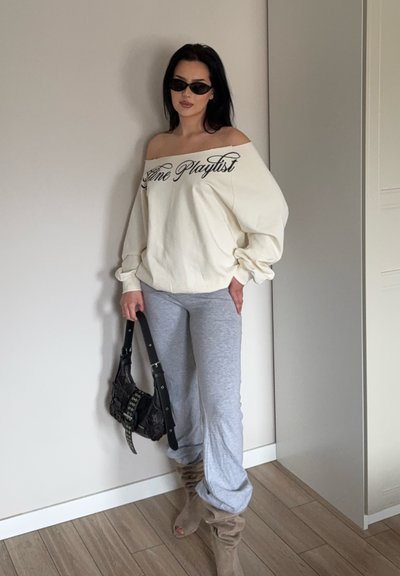 Mujer con sudadera crema con hombros descubiertos y texto "Femme Playlist", pantalones grises, botas beige holgadas, gafas de sol negras, sosteniendo un bolso negro en interiores.
