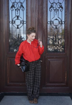 Mujer con chaqueta roja y pantalones de cuadros de pie frente a grandes puertas de madera ornamentadas, sosteniendo gafas de sol y un bolso negro.