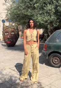 Žuti laneni skraćeni halter top s vezanjem, u kombinaciji s širokim hlačama. U pozadini se nalazi velika skulpturalna glava i zelena kola.