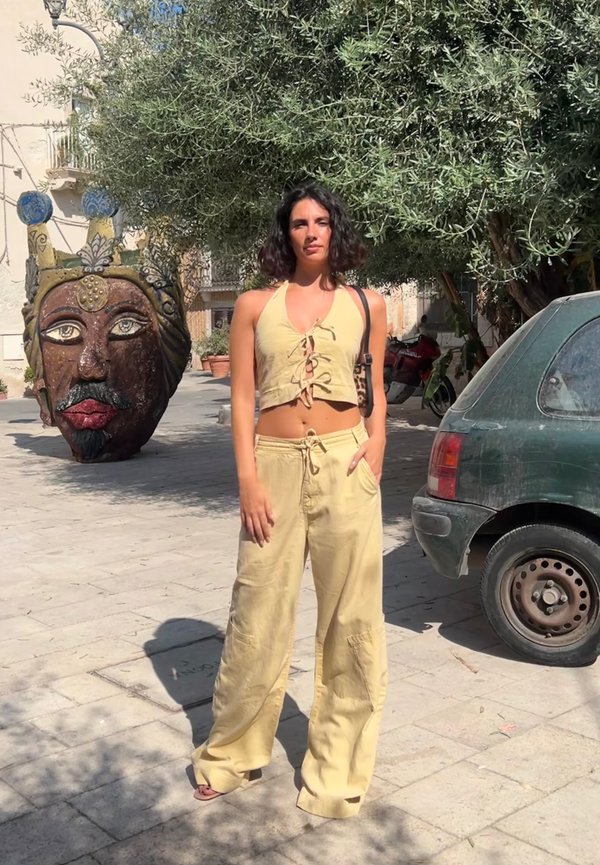 Žuti laneni skraćeni halter top s vezanjem, u kombinaciji s širokim hlačama. U pozadini se nalazi velika skulpturalna glava i zelena kola.