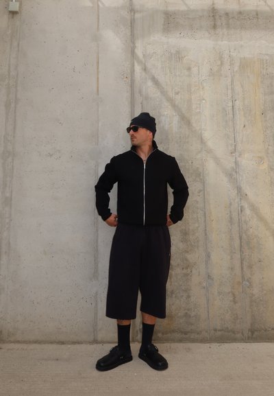 Hombre con gorro negro, gafas de sol, chaqueta con cremallera, pantalones cortos largos, calcetines y zapatos, de pie contra una pared de hormigón con las manos en las caderas.