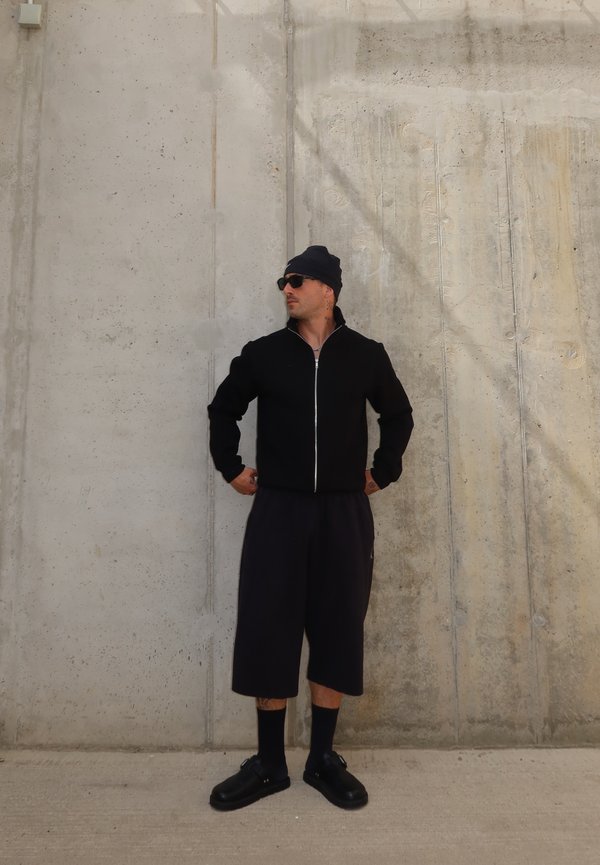 Homme portant un bonnet noir, des lunettes de soleil, une veste zippée, un short long, des chaussettes et des chaussures, debout contre un mur en béton les mains sur les hanches.