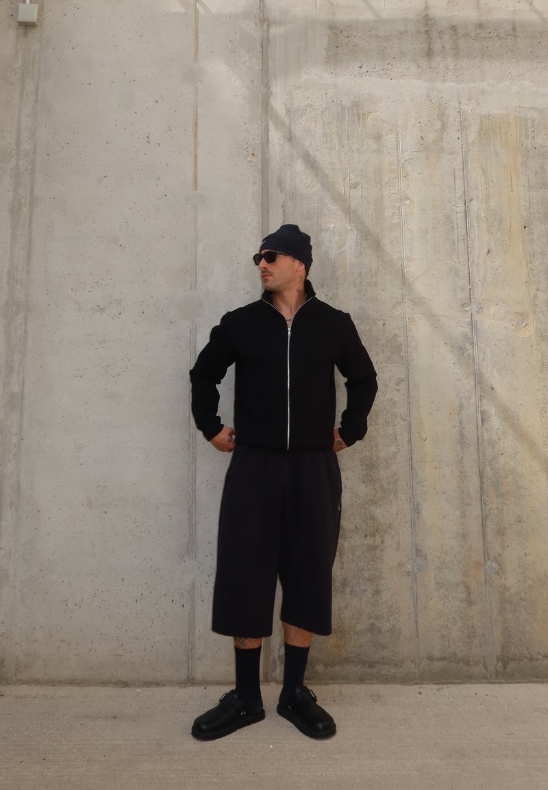 Mann mit schwarzer Mütze, Sonnenbrille, Reißverschlussjacke, langen Shorts, Socken und Schuhen, stehend an einer Betonwand mit Händen in der Hüfte.
