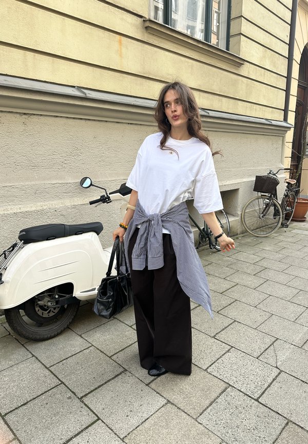 Jeune femme aux cheveux ondulés se tenant sur un trottoir en ville, portant une chemise blanche, un pull rayé noué à la taille, un pantalon large noir, tenant un sac à main noir près d'un scooter blanc.