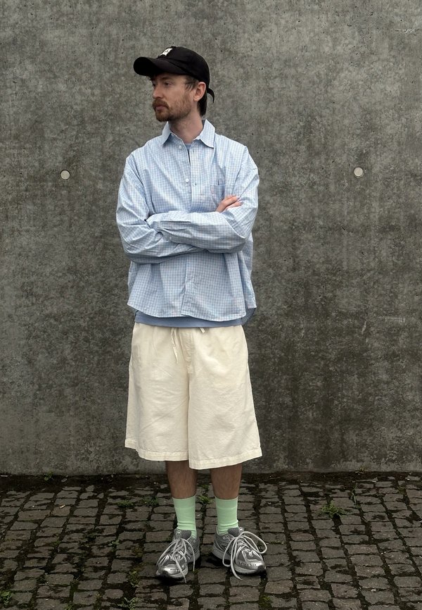 Mand med skæg iført sort kasket, lyseblå ternet skjorte, hvide shorts, grønne sokker og grå sneakers står med armene krydset mod en betonvæg.