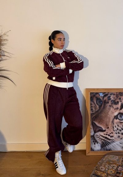 Mujer con chándal Adidas granate y zapatillas blancas apoyada contra la pared, junto a una foto enmarcada de primer plano del rostro de un leopardo.