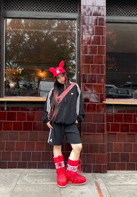 Jaqueta atlética preta com riscas brancas, calções pretos e botas altas vermelhas com marca. Boné vermelho com orelhas e uma mala de ombro estampada.