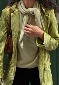 Groene gestructureerde blazer, lichtgroene sjaal en beige top. Materiaal lijkt zacht. Outfit gecombineerd met donkere broek en een bruine handtas.